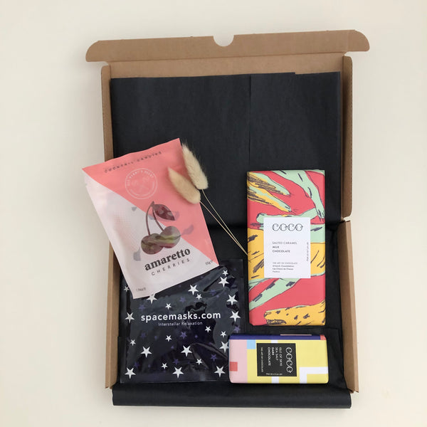 A letterbox friendly gift box containing self heating eye mask from Spacemasks, prosecco gummy bears and a large salted caramel milk chocolate bar and small dark chocolate isle of sea salt bar from Coco Chocolatier on a white background
